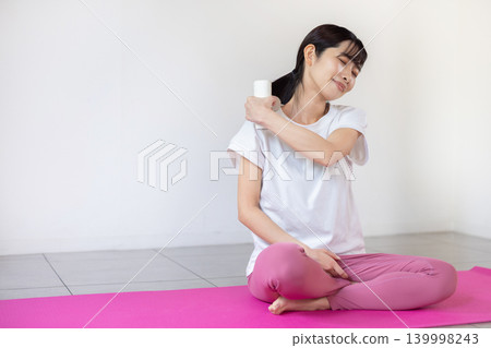 一位年輕的日本女性在瑜珈墊上用按摩槍按摩肩膀。 139998243