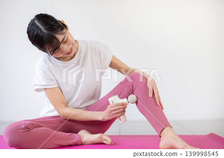 A young Japanese woman self-massages her legs with a massage gun on a yoga mat. 139998545
