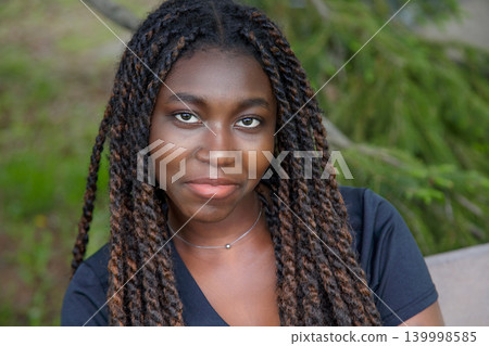 Close-up outdoor portrait of a serene young Black woman with long twists looking at the camera against green nature 139998585
