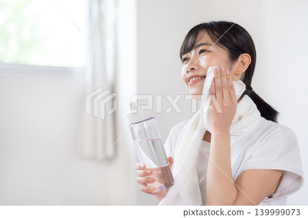 A young Japanese woman smiles in a brightly lit room, holding a water bottle and wiping sweat from her brow with a towel. A young Japanese woman smiles in a brightly lit room, holding a water bottle and wiping sweat from her brow with a towel. 139999073
