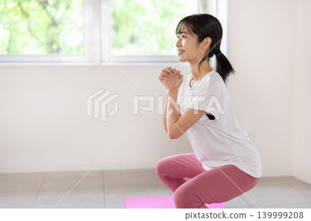 一位年輕的日本女子麵帶微笑，在燈光明亮的房間裡做深蹲。 139999208