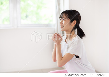 一位年輕的日本女子麵帶微笑，在燈光明亮的房間裡做深蹲。 139999209