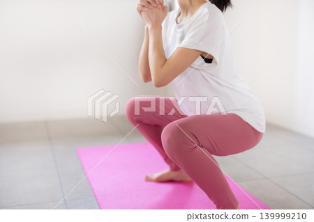 一位年輕的日本女子麵帶微笑，在燈光明亮的房間裡做深蹲。 139999210