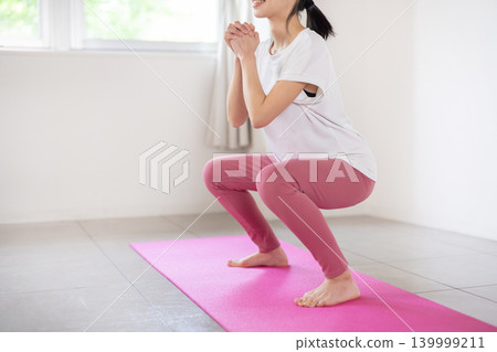 A young Japanese woman smiles and does squats in a brightly lit room. A young Japanese woman smiles and does squats in a brightly lit room. 139999211