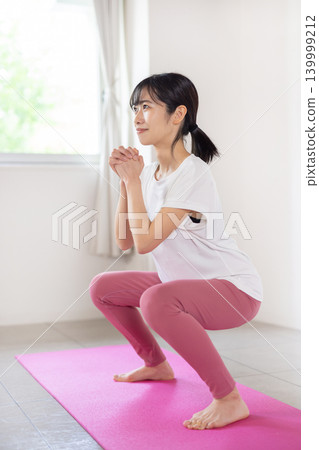 一位年輕的日本女子麵帶微笑，在燈光明亮的房間裡做深蹲。 139999212