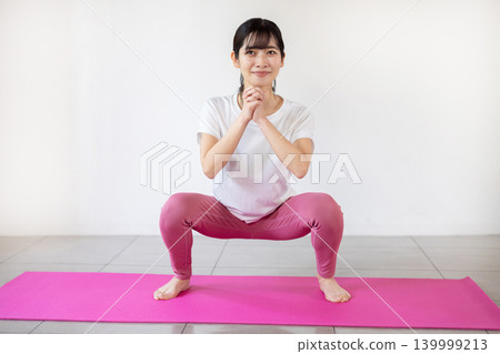A young Japanese woman smiles and does squats in a brightly lit room. A young Japanese woman smiles and does squats in a brightly lit room. 139999213