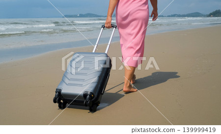 Young woman in pink dress and straw hat rolling a grey suitcase barefoot along sandy beach beside blue sea. Solo vacation concept 139999419