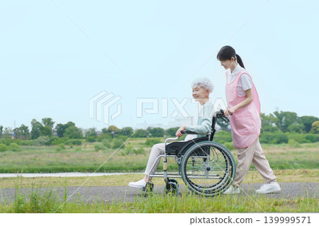 A Japanese senior woman in a wheelchair and a young Japanese female caregiver are strolling along the riverbank. A Japanese senior woman in a wheelchair and a young Japanese female caregiver are strolling along the riverbank. 139999571