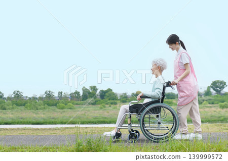 A Japanese senior woman in a wheelchair and a young Japanese female caregiver are strolling along the riverbank. A Japanese senior woman in a wheelchair and a young Japanese female caregiver are strolling along the riverbank. 139999572