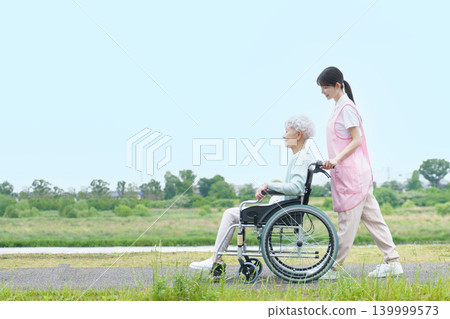 一位坐在輪椅上的日本老婦和一位年輕的日本女護理員正在河岸邊散步。 139999573