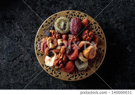 Dried tropical fruits, nuts, and raisins in a golden bowl on black stone table. Dried tropical fruits, nuts, and raisins in a golden bowl on black stone table. 140000031