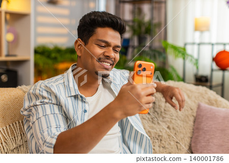 Indian man on home sofa smiling at smartphone message in living room and enjoying cheerful good news Indian man on home sofa smiling at smartphone message in living room and enjoying cheerful good news 140001786