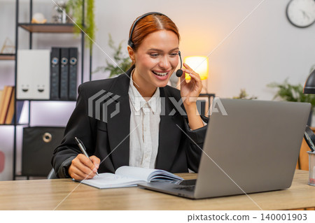 Young businesswoman wears headset and takes notes during client video call on laptop smiling calmly 140001903