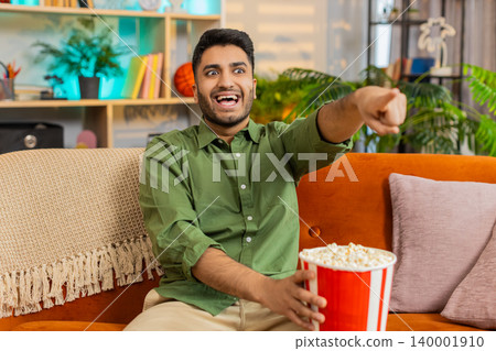 Indian man on home sofa watching movie with popcorn laughing and pointing during fun film night joy Indian man on home sofa watching movie with popcorn laughing and pointing during fun film night joy 140001910