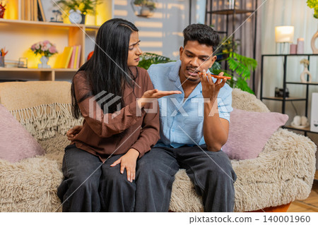 Indian couple man and woman sitting on home sofa arguing by smartphone during complaint call at home Indian couple man and woman sitting on home sofa arguing by smartphone during complaint call at home 140001960