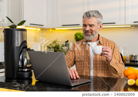 Middle-aged man working on laptop in kitchen sipping coffee smiling during morning daily job at home 140002009