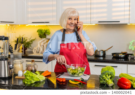 Senior old woman mixing vegetable salad while talking on phone in home kitchen for healthy diet plan Senior old woman mixing vegetable salad while talking on phone in home kitchen for healthy diet plan 140002041
