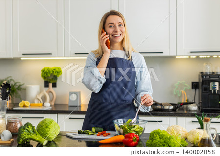 Young woman talking on orange smartphone while mixing salad in kitchen sharing healthy keto recipe Young woman talking on orange smartphone while mixing salad in kitchen sharing healthy keto recipe 140002058