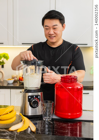 Asian man using blender to mix banana milk cottage cheese whey protein shake in home kitchen daily Asian man using blender to mix banana milk cottage cheese whey protein shake in home kitchen daily 140002356