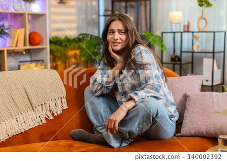 Young woman on home sofa suffering tooth pain after drinking water and feeling worse at home alone Young woman on home sofa suffering tooth pain after drinking water and feeling worse at home alone 140002392
