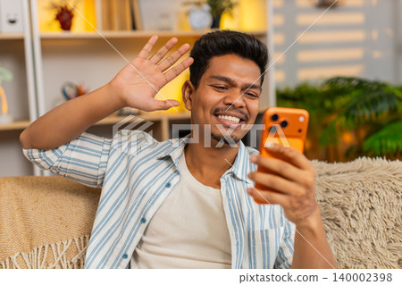 Indian man on home sofa waving at smartphone camera during video call greeting and farewell at home 140002398
