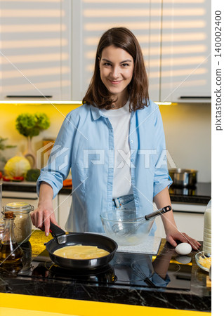 Young woman cooking cheese omelet pouring egg mix into frying pan in home kitchen for healthy diet 140002400