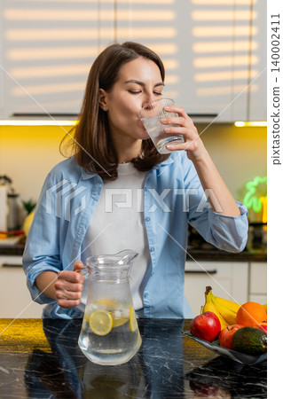 Young woman pouring cold lemon water in home kitchen to quench thirst for diet hydration fit routine 140002411