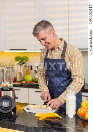 Middle-aged man slicing banana for blender in kitchen preparing smoothie for diet nutrition routine Middle-aged man slicing banana for blender in kitchen preparing smoothie for diet nutrition routine 140002417