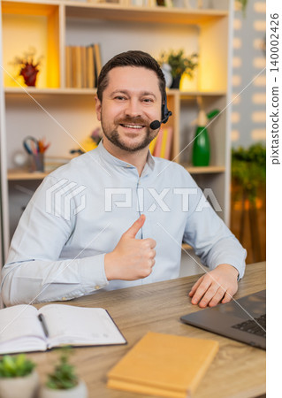 Young man on home table working in headset with laptop and smiling during virtual customer support 140002426