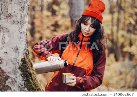 Woman pours liquid from thermos into cup while standing next to tree in autumn forest. reusable stainless thermos, zero waste lifestyle, sustainable drinking habits, plastic free alternative. Woman pours liquid from thermos into cup while standing next to tree in autumn forest. reusable stainless thermos, zero waste lifestyle, sustainable drinking habits, plastic free alternative. 140002667