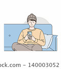 A young man wearing a beanie sits cross-legged on a blue couch, absorbed in his smartphone, with a pillow nearby. 140003052