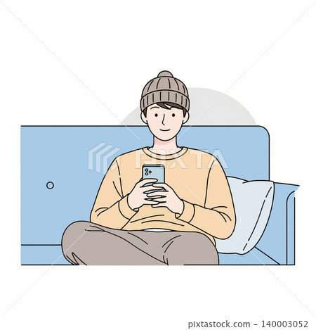 A young man wearing a beanie sits cross-legged on a blue couch, absorbed in his smartphone, with a pillow nearby. A young man wearing a beanie sits cross-legged on a blue couch, absorbed in his smartphone, with a pillow nearby. 140003052