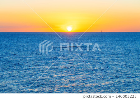 A tranquil scene unfolds as the sun ascends above the deep blue sea, casting a warm glow across the water and sky, with a lone sailboat visible in the distance A tranquil scene unfolds as the sun ascends above the deep blue sea, casting a warm glow across the water and sky, with a lone sailboat visible in the distance 140003225