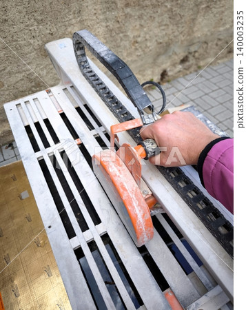 The hand of a worker in a pink jacket guides the cutting blade of a water-cooled electric tile cutter. The hand of a worker in a pink jacket guides the cutting blade of a water-cooled electric tile cutter. 140003235