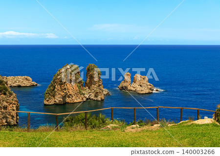 A picturesque summer landscape featuring dramatic rock formations emerging from the deep blue ocean, bordered by a vibrant green grassy area and a rustic wooden fence A picturesque summer landscape featuring dramatic rock formations emerging from the deep blue ocean, bordered by a vibrant green grassy area and a rustic wooden fence 140003266
