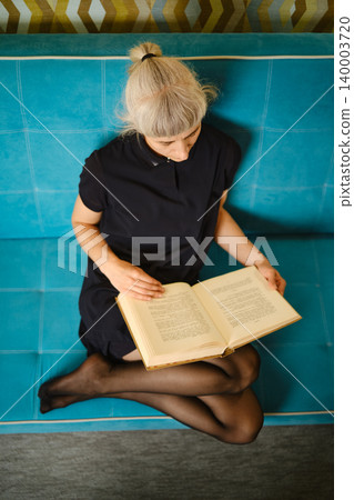 Person sits on couch and reads book in casual indoor setting during day Person sits on couch and reads book in casual indoor setting during day 140003720
