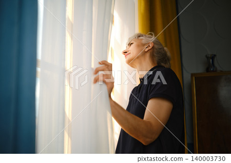 Woman looking out the window in morning light with sheer curtains drawn aside Woman looking out the window in morning light with sheer curtains drawn aside 140003730