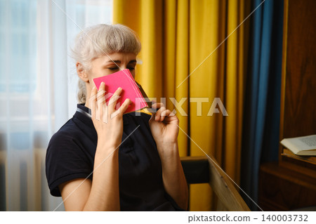 Woman holds a pink book to her face while sitting in a room with yellow and blue curtains Woman holds a pink book to her face while sitting in a room with yellow and blue curtains 140003732