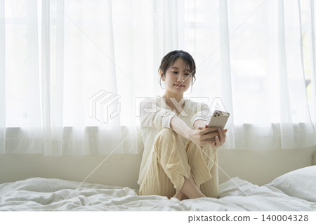 一位年輕女子坐在床上看著她的智慧型手機螢幕。 140004328