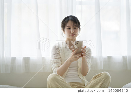 一位年輕女子坐在床上看著她的智慧型手機螢幕。 140004340