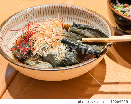 Japanese-style simmered eggplant. Eggplant simmered in a flavorful broth. Topped with bonito flakes. The eggplant is being lifted with chopsticks. Japanese-style simmered eggplant. Eggplant simmered in a flavorful broth. Topped with bonito flakes. The eggplant is being lifted with chopsticks. 140004619