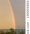 A bright vibrant rainbow stretching down through a moody sky towards coastal trees and the sea. 140004860
