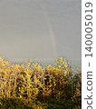 A faint rainbow appearing in the moody sky over sunlit autumn trees near the sea after a storm. 140005019