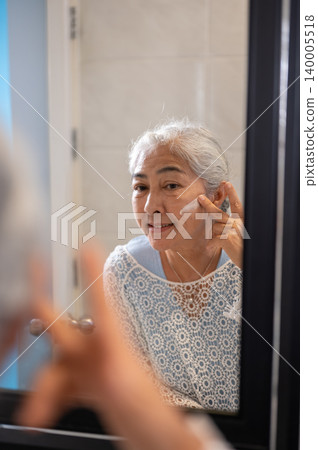 Happy elderly old asian woman grandma applying cream on face looking at bathroom mirror's reflection Happy elderly old asian woman grandma applying cream on face looking at bathroom mirror's reflection 140005518