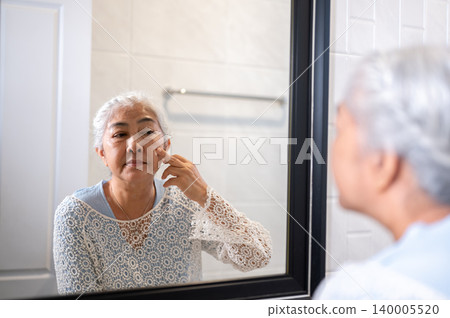 Elderly old asian woman grandma applying cream lotion on face as looking in bathroom mirror at home. Elderly old asian woman grandma applying cream lotion on face as looking in bathroom mirror at home. 140005520