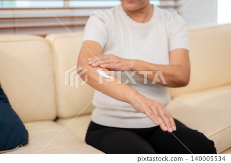 Close up of elderly old asian woman grandma applying skin lotion cream on arm while sitting on sofa. Close up of elderly old asian woman grandma applying skin lotion cream on arm while sitting on sofa. 140005534