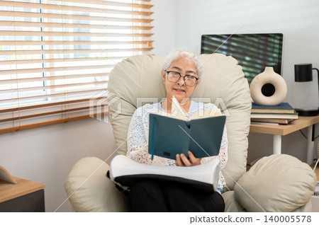 Happy retired elderly old asian woman reading a book on cosy sofa armchair in a living room at home 140005578