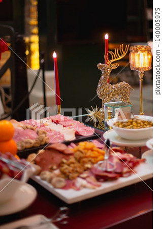 Festive buffet table with charcuterie, dried fruit, nuts, olives, and a crystal reindeer figurine flanked by red candles at an elegant evening event. Festive buffet table with charcuterie, dried fruit, nuts, olives, and a crystal reindeer figurine flanked by red candles at an elegant evening event. 140005975