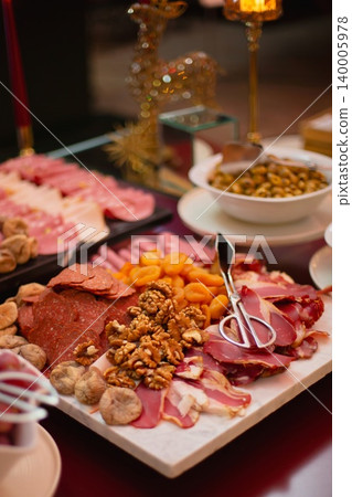 Charcuterie board loaded with sliced salami, cured meats, walnuts, dried figs, and apricots, served with tongs at an elegant buffet. Charcuterie board loaded with sliced salami, cured meats, walnuts, dried figs, and apricots, served with tongs at an elegant buffet. 140005978