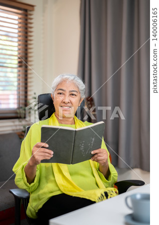 Happy elderly old asian woman thinking while holding a book sitting in living room at home or office Happy elderly old asian woman thinking while holding a book sitting in living room at home or office 140006165